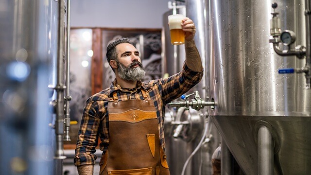 Smiling brewer holding a glass of beer in a brewery, surrounded by brewing equipment.