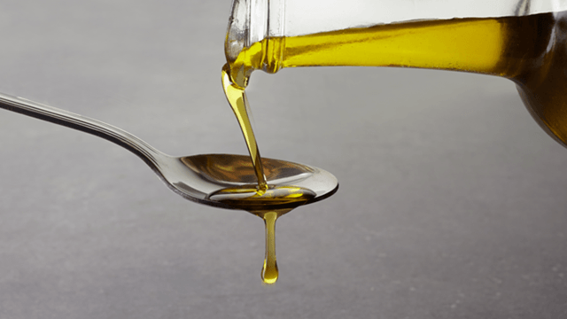 Olive oil being poured from a bottle into a metal spoon, with some oil dripping off the spoon
