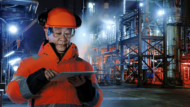 male Asian engineer wearing hard hat and safety glasses holding ipad infront of industrial plant