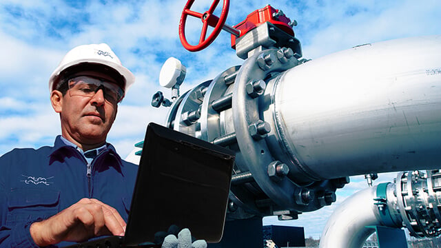 engineer wearing hard hat and safety glasses holding laptop in front of pipes and blue sky