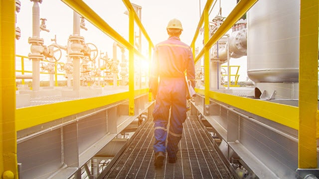 Engineer wearing hard hat seen from behind walking into the setting sun at industrial plant